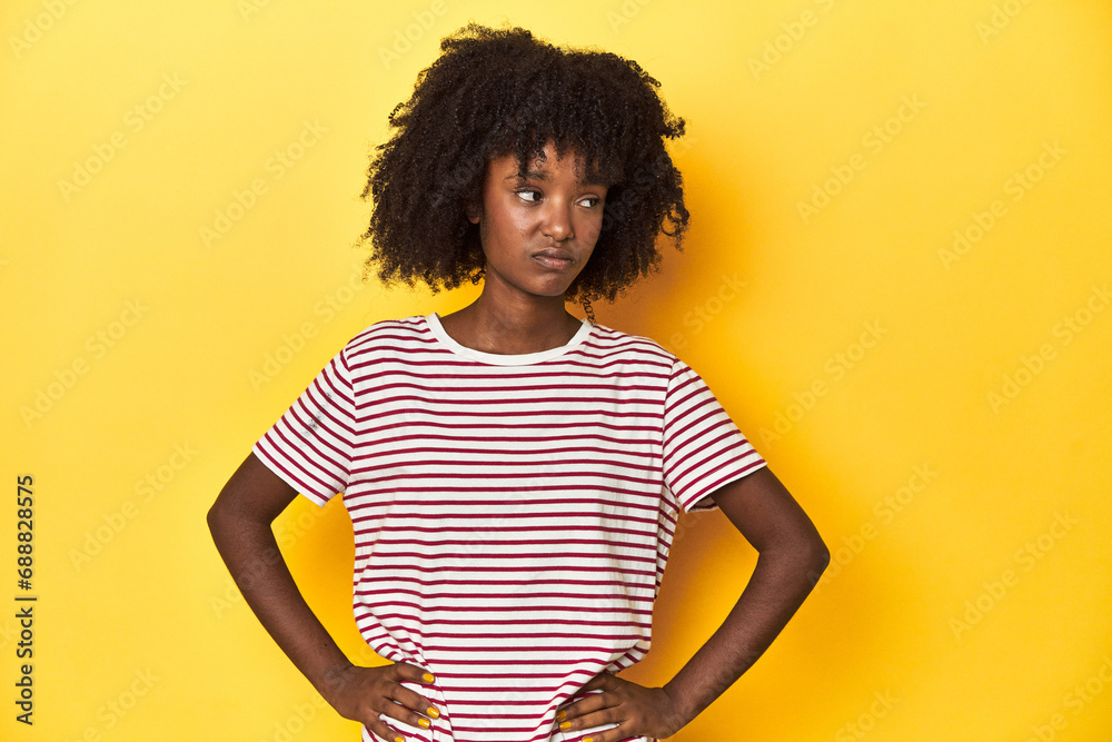 Teen girl in red striped T-shirt, yellow studio backdrop confused, feels doubtful and unsure.