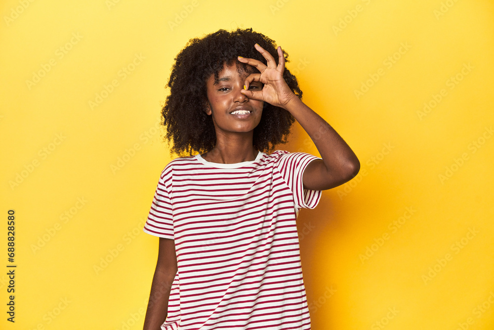 Teen girl in red striped T-shirt, yellow studio backdrop excited keeping ok gesture on eye. Teen girl in red striped T-shirt, yellow studio backdrop excited keeping ok gesture on eye.