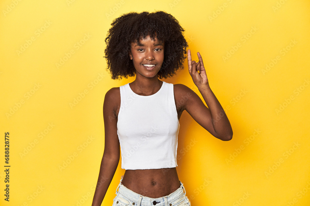Teen girl in white tank top, yellow studio background showing a horns gesture as a revolution concept. Teen girl in white tank top, yellow studio background showing a horns gesture as a revolution concept.