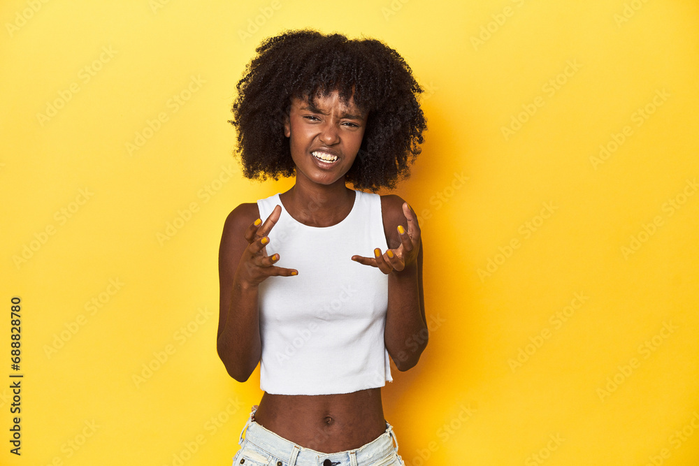 Teen girl in white tank top, yellow studio background upset screaming with tense hands.