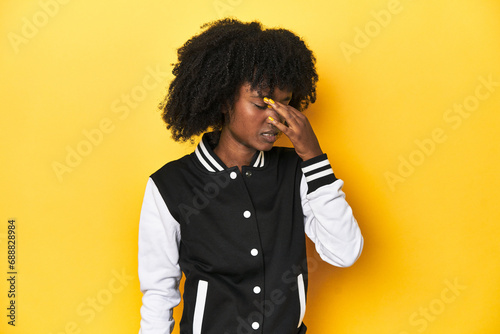 Teen girl in baseball jacket, yellow studio background having a head ache, touching front of the face. photo