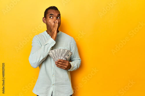 African American man with dollar bills, yellow studio, is saying a secret hot braking news and looking aside