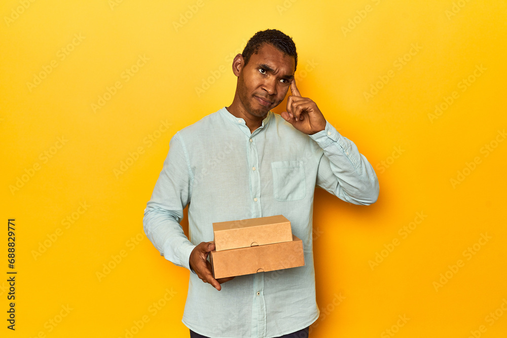 African American man with food boxes, yellow studio, pointing temple with finger, thinking, focused on a task. African American man with food boxes, yellow studio, pointing temple with finger, thinking, focused on a task.
