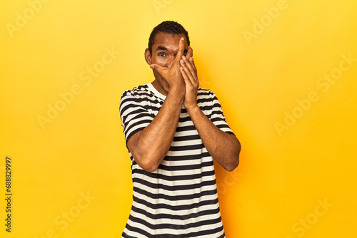 African American man in striped t-shirt, yellow studio, blink through fingers frightened and nervous.