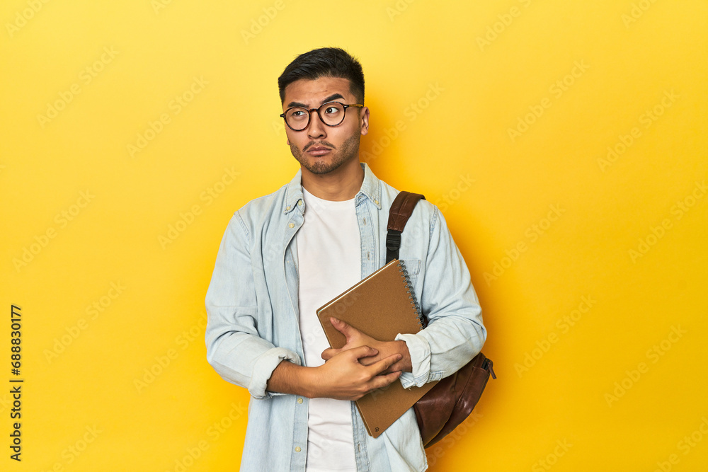 Asian student with backpack and notebook, yellow studio backdrop confused, feels doubtful and unsure. Asian student with backpack and notebook, yellow studio backdrop confused, feels doubtful and unsure.