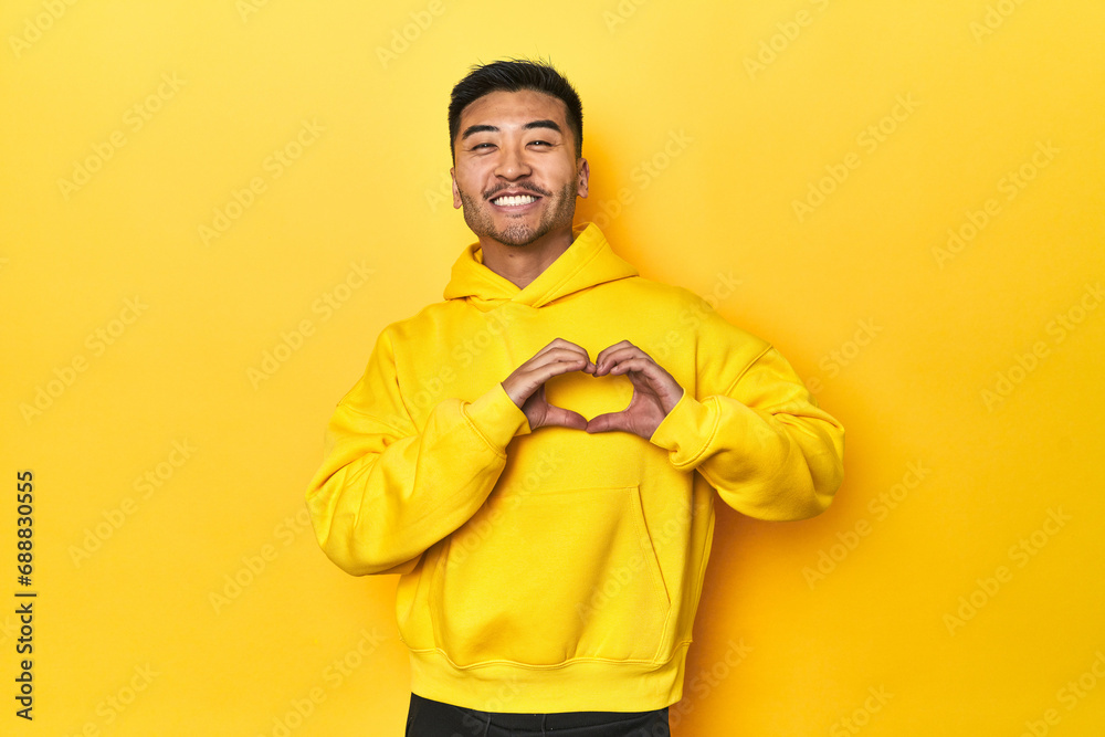 Bold Asian man in yellow hoodie on yellow studio smiling and showing a heart shape with hands. Bold Asian man in yellow hoodie on yellow studio smiling and showing a heart shape with hands.