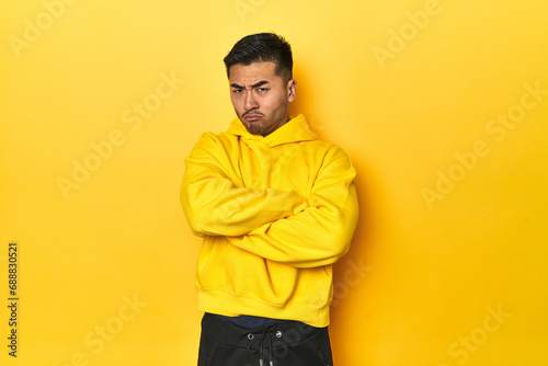 Bold Asian man in yellow hoodie on yellow studio frowning face in displeasure, keeps arms folded.