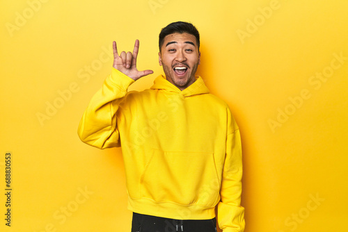 Bold Asian man in yellow hoodie on yellow studio showing a horns gesture as a revolution concept.