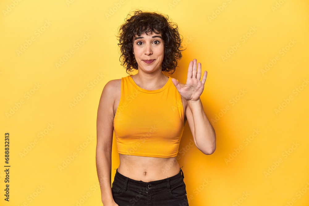 Curly-haired Caucasian woman in yellow top smiling cheerful showing number five with fingers. Curly-haired Caucasian woman in yellow top smiling cheerful showing number five with fingers.