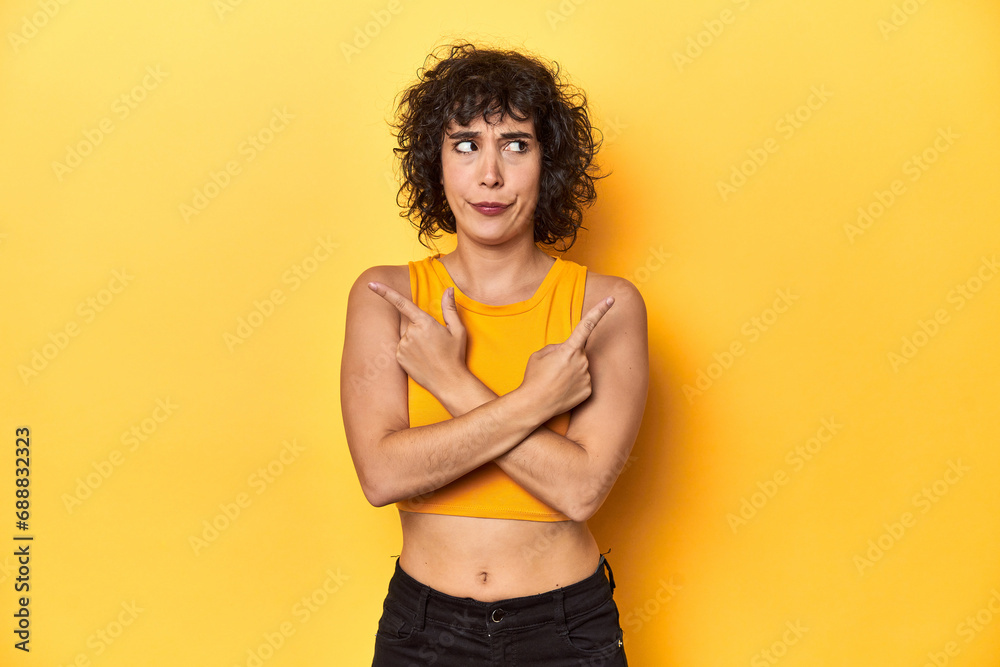 Curly-haired Caucasian woman in yellow top points sideways, is trying to choose between two options.