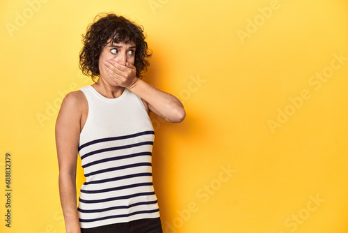 Caucasian curly-haired woman in white tank-top thoughtful looking to a copy space covering mouth with hand.