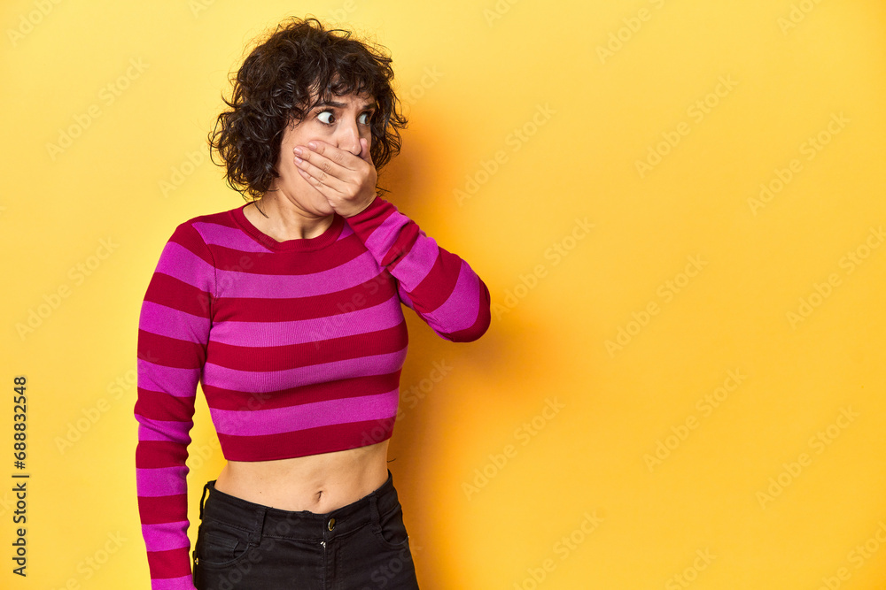 Caucasian curly-haired woman in pink striped-top thoughtful looking to a copy space covering mouth with hand.