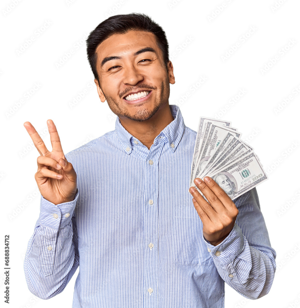Young Chinese man holding dollar bills in studio joyful and carefree showing a peace symbol with fingers.