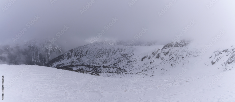 Obraz premium The white snow peaks of Pirin partly covered by fog. Snowy weather conditions for winter sports and tourism. Alpine ski resort Bansko, Bulgaria.