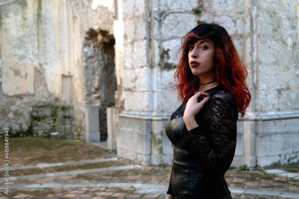 Amidst ruins, a red-haired dark beauty, half-bust framed, explores the mystery of an abandoned abbey with a haunting allure.