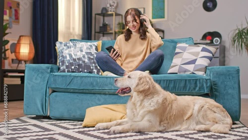 Beautiful Caucasian woman with headphones comfortably sitting in lotus position on couch in cozy room. Young female holding phone and listening music while her cute dog laying on carpet. photo