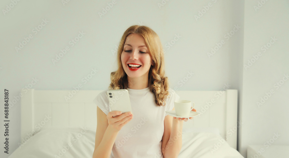 Happy smiling young woman with mobile phone holding cup of coffee in bed in white bedroom at home in the morning Happy smiling young woman with mobile phone holding cup of coffee in bed in white bedroom at home in the morning
