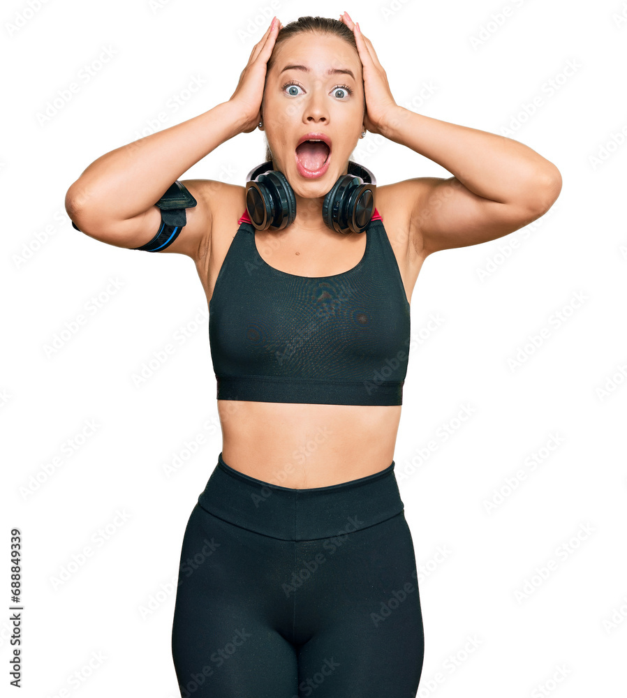 Beautiful blonde woman wearing gym clothes and using headphones crazy and scared with hands on head, afraid and surprised of shock with open mouth