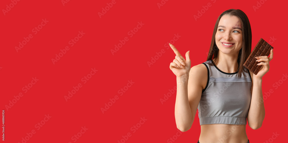 Happy young woman holding chocolate bar and pointing at something on red background with space for text Happy young woman holding chocolate bar and pointing at something on red background with space for text