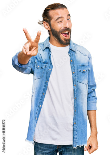 Attractive man with long hair and beard wearing casual denim jacket smiling with happy face winking at the camera doing victory sign with fingers. number two.