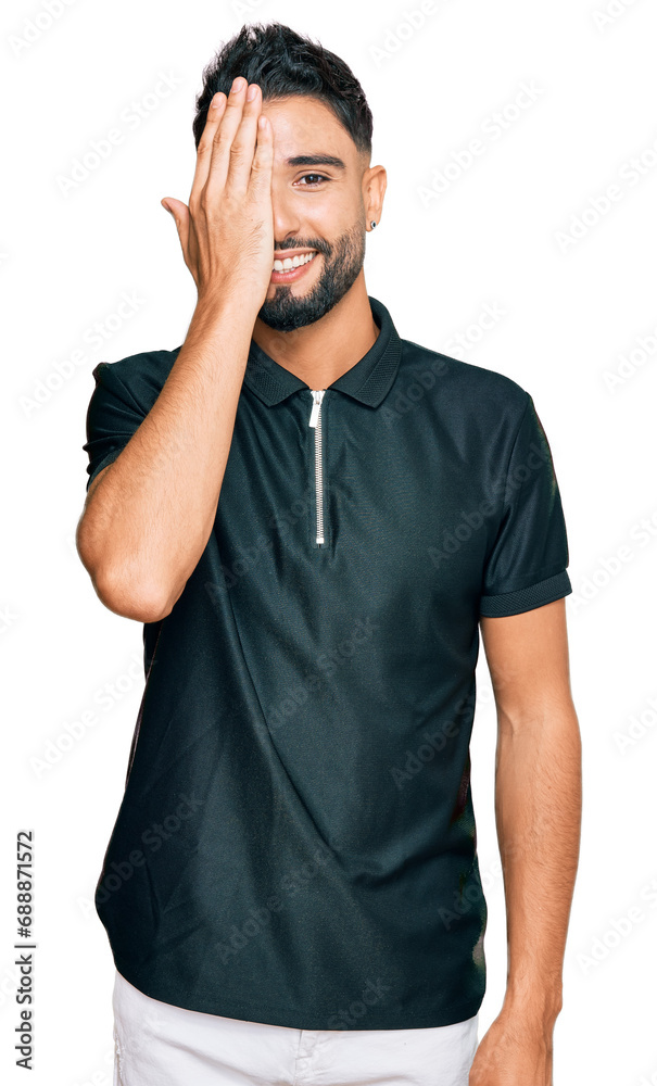 Young man with beard wearing sportswear covering one eye with hand, confident smile on face and surprise emotion. Young man with beard wearing sportswear covering one eye with hand, confident smile on face and surprise emotion.