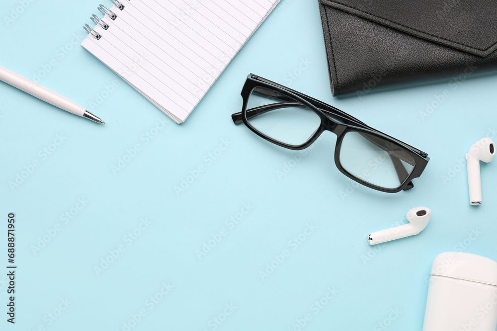 Composition with stylish eyeglasses, modern earphones, notebook and case on blue background Composition with stylish eyeglasses, modern earphones, notebook and case on blue background
