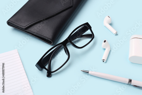 Composition with stylish eyeglasses, modern earphones, notebook and case on blue background, closeup