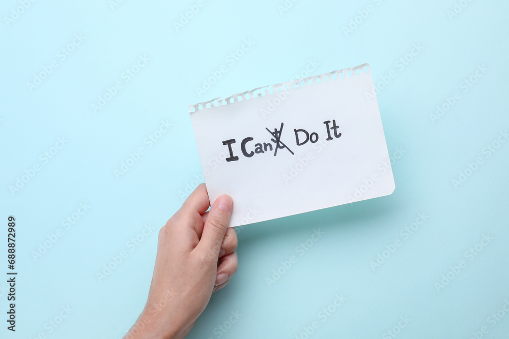 Motivation concept. Woman holding paper with changed phrase from I Can't Do It into I Can Do It by crossing over letter T on light blue background, top view Motivation concept. Woman holding paper with changed phrase from I Can't Do It into I Can Do It by crossing over letter T on light blue background, top view