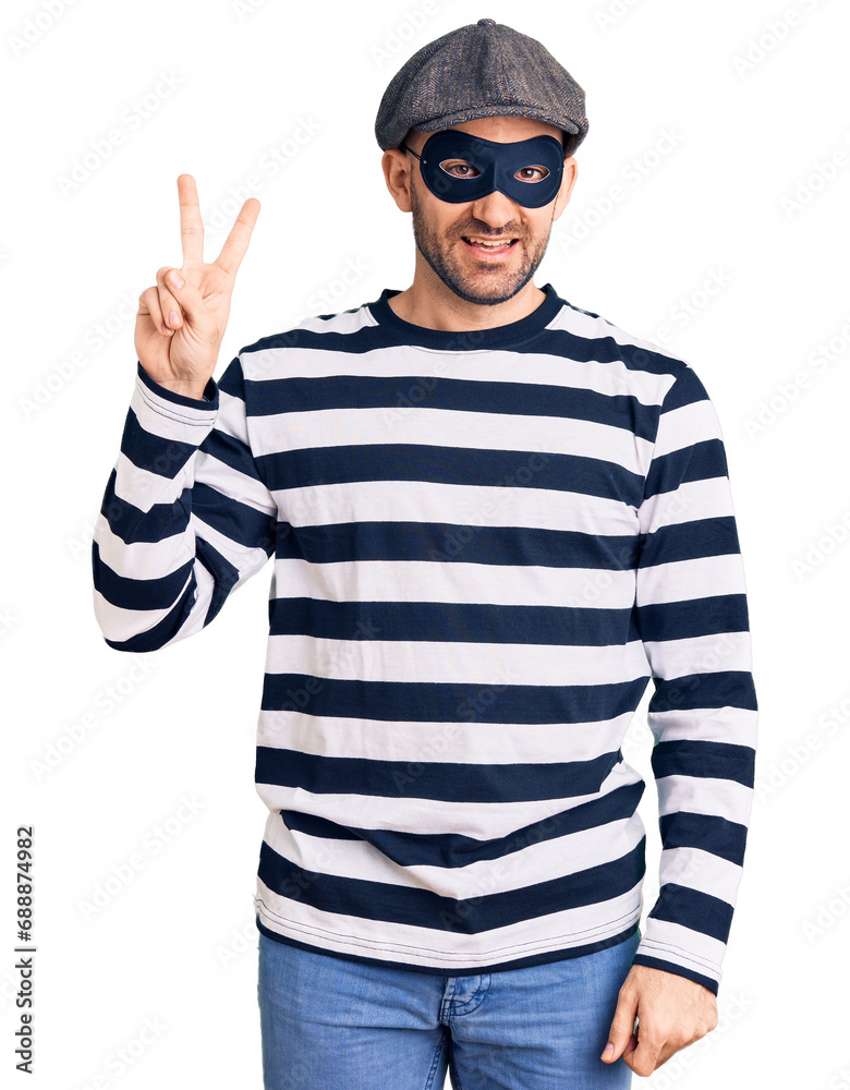 Young handsome man wearing burglar mask smiling looking to the camera showing fingers doing victory sign. number two. Young handsome man wearing burglar mask smiling looking to the camera showing fingers doing victory sign. number two.