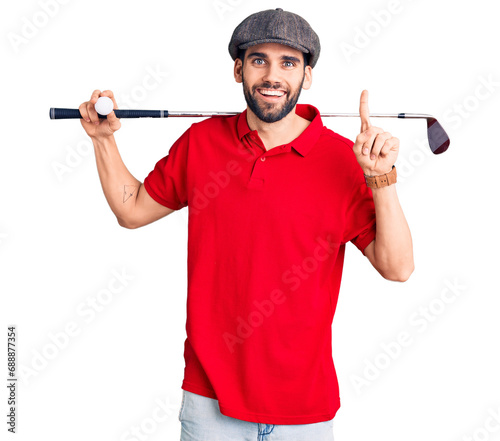 Young handsome man with beard playing golf holding club and ball surprised with an idea or question pointing finger with happy face, number one