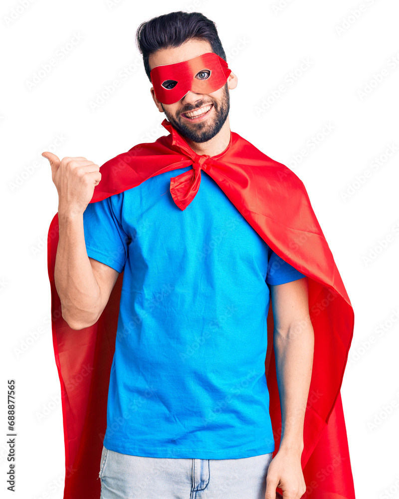 Young handsome man with beard wearing super hero costume smiling with happy face looking and pointing to the side with thumb up. Young handsome man with beard wearing super hero costume smiling with happy face looking and pointing to the side with thumb up.