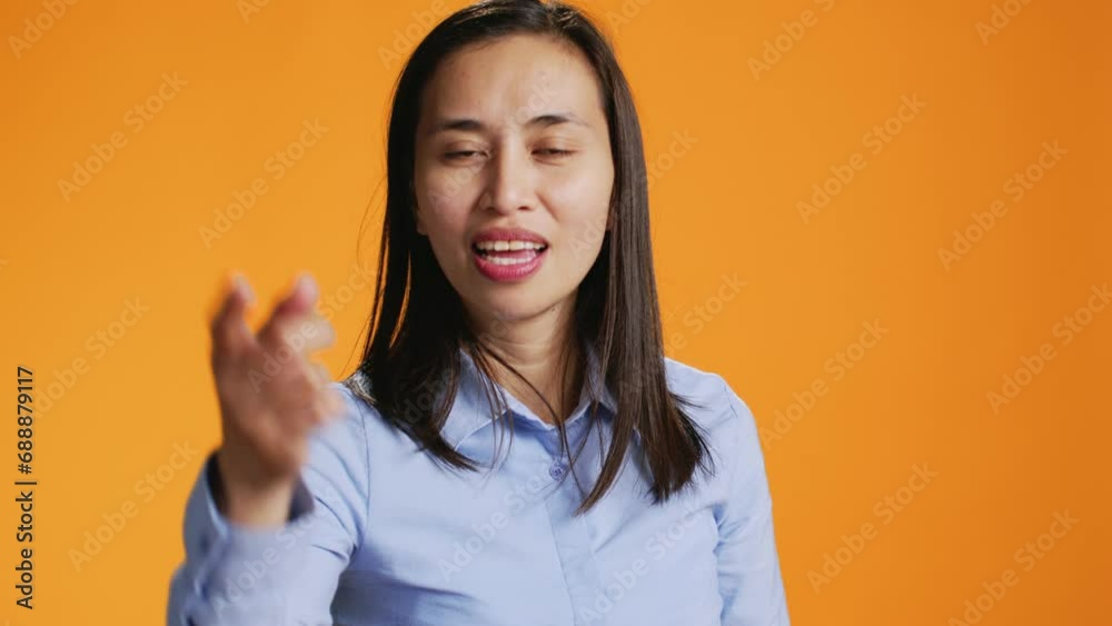 Happy asian person pointing finger at camera during shoot time. Positive young woman with casual attitude using charisma, standing in studio over orange background, showing something.