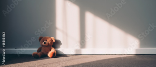 Lonely forgotten stuffed bear toy sitting against an empty room with morning sunlight and shadows against the wall, sad broken spirit waiting for someone to pick him up and take to a new home. photo