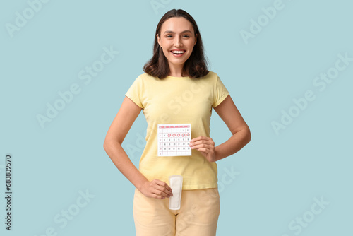 Beautiful young woman holding pad and calendar with marked days of menstruation on blue background