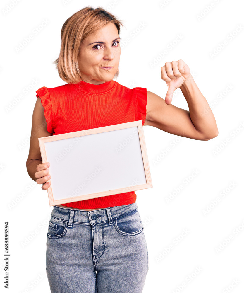 Young blonde woman holding empty white chalkboard with angry face, negative sign showing dislike with thumbs down, rejection concept