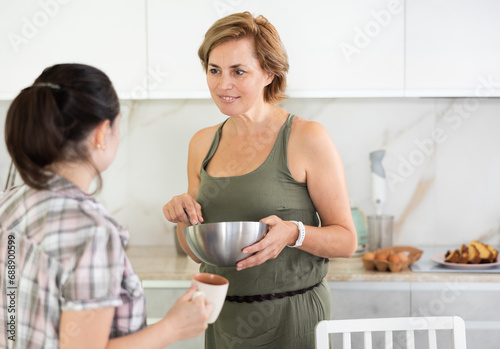 Two women are cooking lunch and chatting nicely in kitchen. Middle-aged woman is stirring pancake batter in bowl and listening to female friend story of relationship with boyfriend