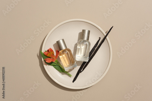 On a gray background, two glass bottles displayed with red flower and vanilla sticks on a round ceramic dish. Minimal concept for advertising, mockup for design