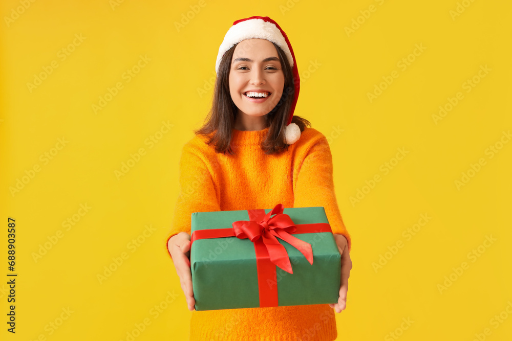 Happy young woman in Santa hat with Christmas gift box on yellow background Happy young woman in Santa hat with Christmas gift box on yellow background