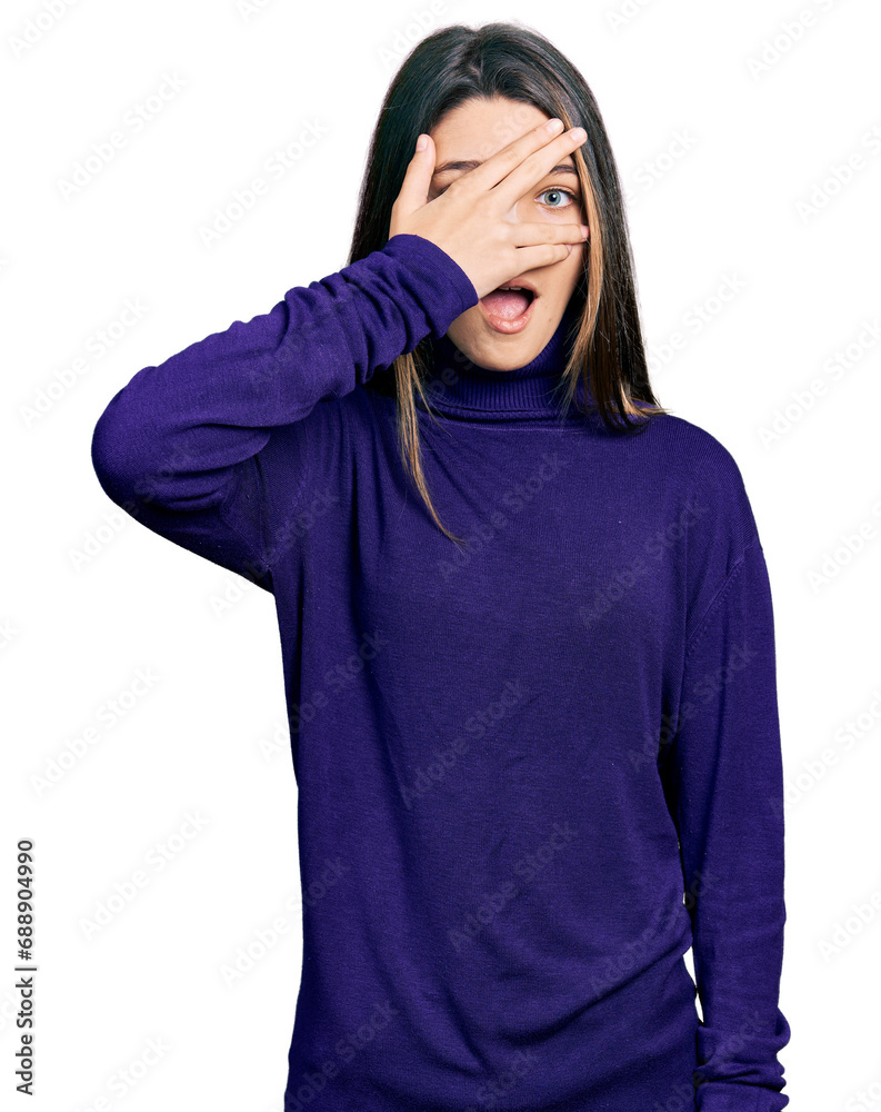 Young brunette girl with long hair wearing turtleneck sweater peeking in shock covering face and eyes with hand, looking through fingers with embarrassed expression.