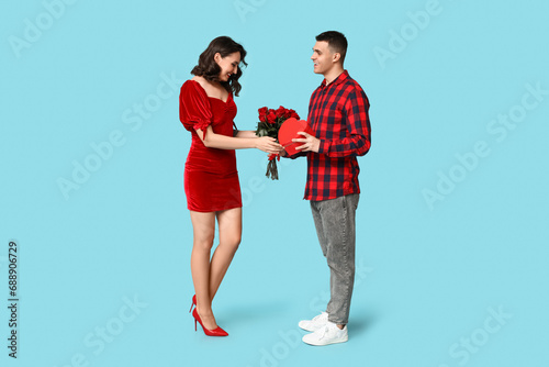Loving young couple with gift box and bouquet of beautiful roses on blue background. Celebration of Saint Valentine's Day