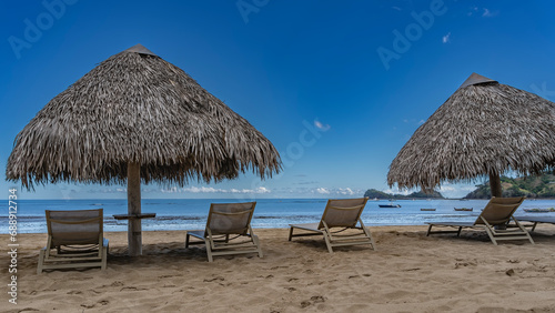 Sunbeds stand in a row on the sandy beach in the shade of straw sun umbrellas. Boats in the calm blue ocean. Light clouds in the azure sky. Islands in the distance. Madagascar. Nosy Be  