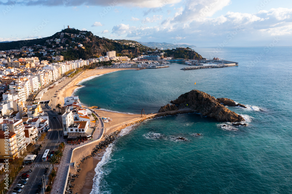 Scenic drone view of coastal Catalan town of Blanes overlooking white residential buildings and Sa Palomera Rock on background of San Juan hill with ancient castle on sunny day, Barcelona, Spain ..