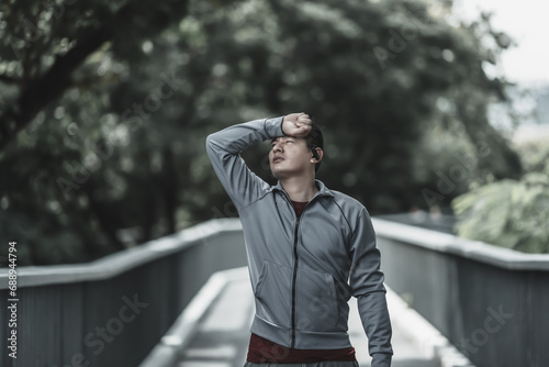 a tired young man wipe the sweat. Man resting, breathing and holding a bottle of drinking water on the road in the park morning. Health and Lifestyle in big city life concept. photo