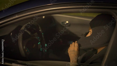 a black man sitting in the drivers seat of a car in broad daylight pulls a black ski mask and hood over his head then picks up a handgun and prepares to load a bullet in the chamber photo