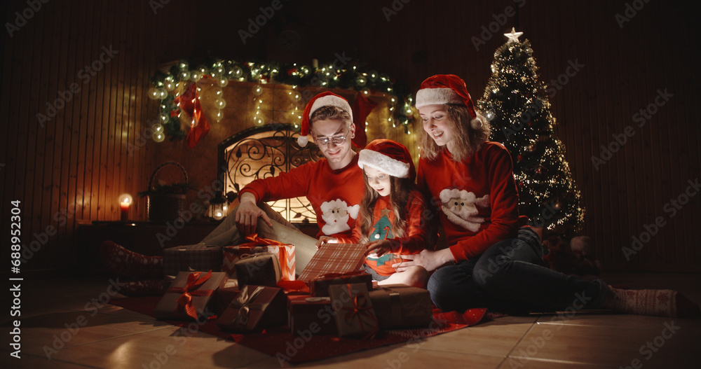 Caucasian family of three sitting in decorated room near christmas tree, little girl opening her christmas gift with something special - christmas spirit, togetherness concept