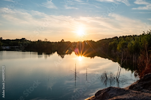 This image captures the serene atmosphere of a quarry lake at sunset. The sun hovers just above the treeline, casting a soft glow over the still water. The lake, flanked by rocky edges and sparse photo