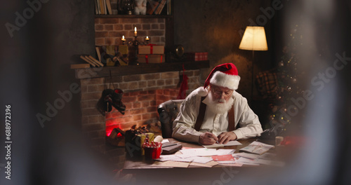 Busy Santa Claus writing notes  working at night  outside view through window. St Nicholas making up checklist of children wishes and presents for kids. Late work in evening on Christmas Eve.