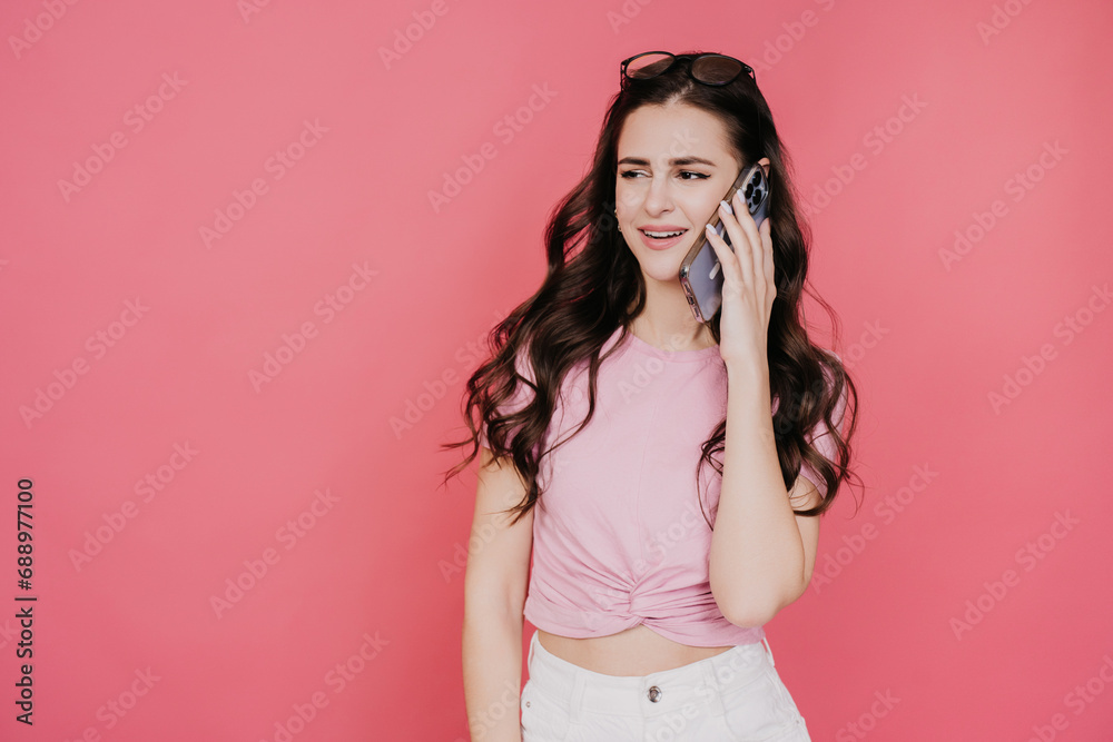 Satisfied brunette girl talking by phone toothy smiling standing against pink studio background looks at copy space. Promo, sale, gossip concept. Satisfied brunette girl talking by phone toothy smiling standing against pink studio background looks at copy space. Promo, sale, gossip concept.