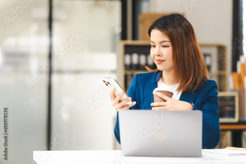 Happy attractive middle aged Asian people advertising manager business woman in formal suit smiles while using laptop, tablet, mobile phone, with laptop and coffee on desk.