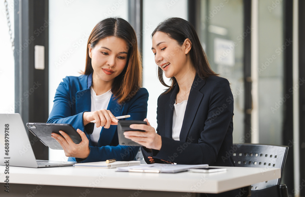 Two middle aged and young Asian female executives in formal suits review bar chart, discussing business strategies in office setting, senior executives or directors in advertising or public relations